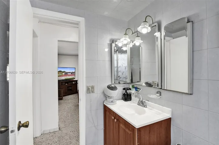Vanity with a solid surface countertop, integrated sink, wooden cabinetry, wall-mounted mirror, and multi-bulb light fixture, finished with full-height tile wall surfacing