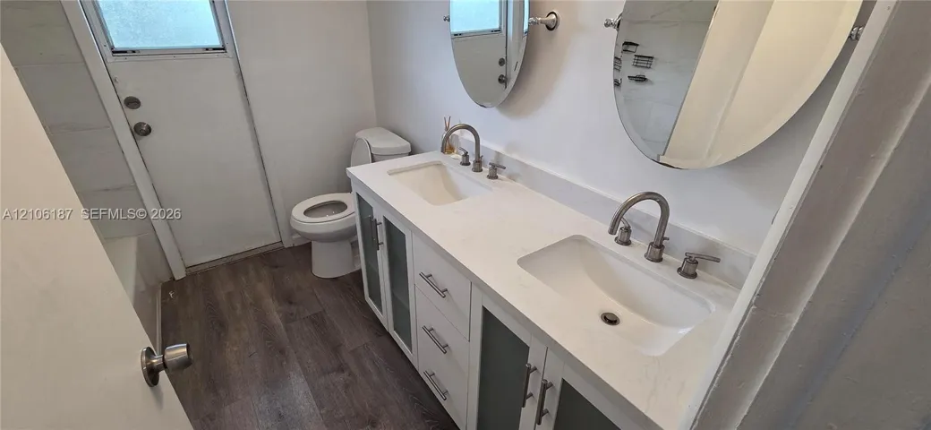 Dual-sink vanity with a solid surface countertop, undermount basins, brushed metal hardware, and frosted glass cabinet inserts, complemented by wood-finish flooring and oval wall-mounted mirrors