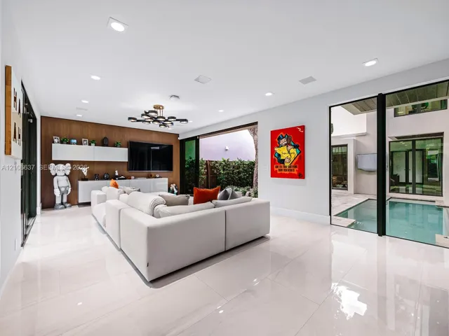 Large-format tile flooring, wood-panel accent wall with integrated built-in cabinetry, recessed ceiling lighting, statement multi-arm chandelier, and floor-to-ceiling glass doors opening to an internal pool area
