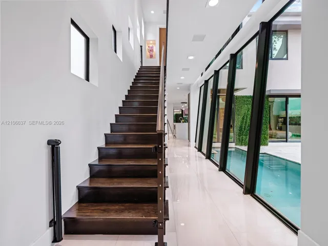 Staircase with dark wood-finish treads and metal railings, large-format tile flooring, recessed ceiling lighting, and floor-to-ceiling black-framed windows overlooking a pool area
