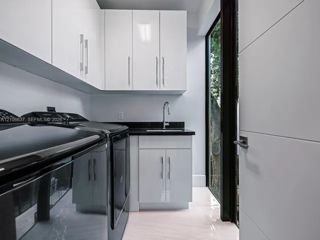Glossy white cabinetry with vertical pulls, solid-surface dark countertop, undermount sink with gooseneck faucet, light-toned polished flooring, and full-height glass paneled door
