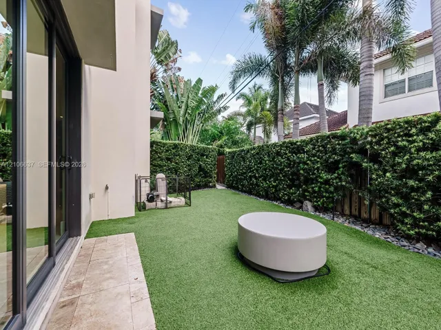 Exterior space featuring an artificial turf lawn, stone-finish patio pavers, perimeter hedges, and sliding glass doors