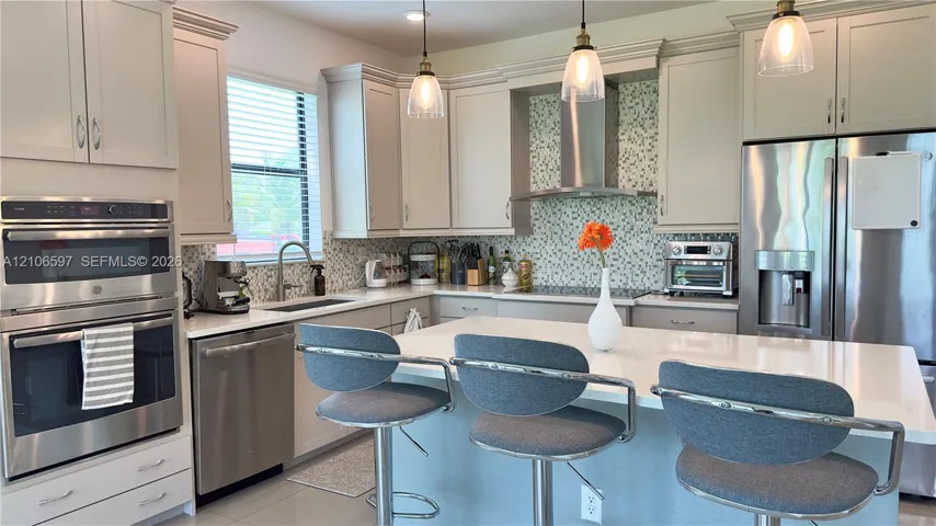 Kitchen featuring light-toned shaker cabinetry, stone-surface countertops, a mosaic tile backsplash, stainless steel built-in double ovens, and a central island with pendant lighting