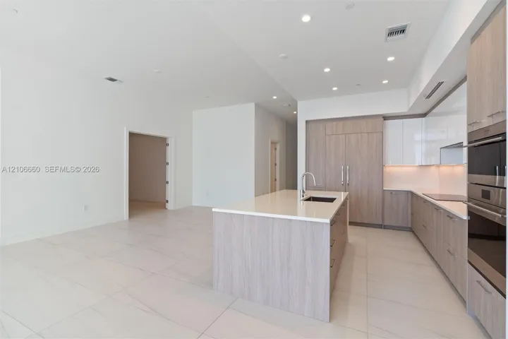 Kitchen featuring wood-finish cabinetry, white high-gloss upper cabinets, solid-surface countertops, large kitchen island with integrated sink, and built-in stainless steel double ovens