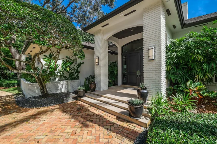 Covered entryway featuring white brick pillars, a multi-paneled dark wood front door with arched transom, tiered concrete steps, and a herringbone-patterned brick paver walkway