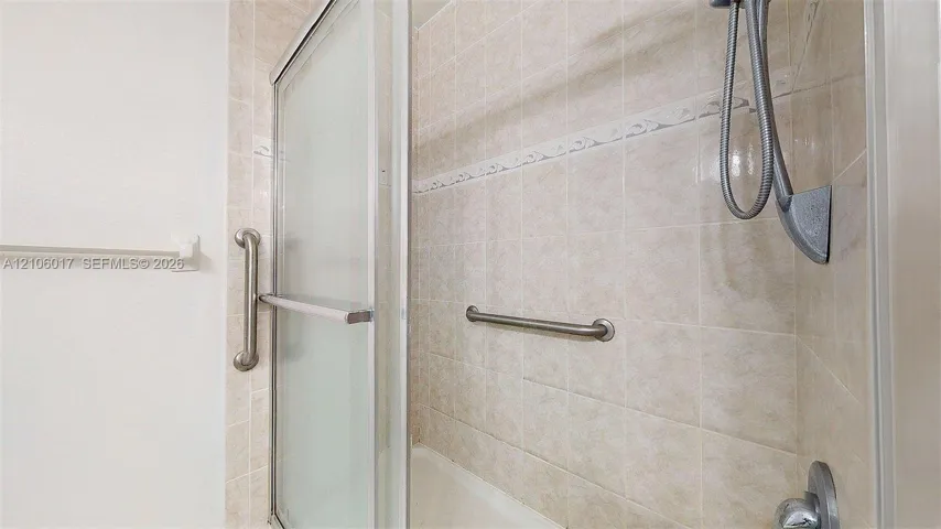 Shower area featuring tiled walls with decorative accents, sliding glass door, handheld shower head, and wall-mounted metal grab bars