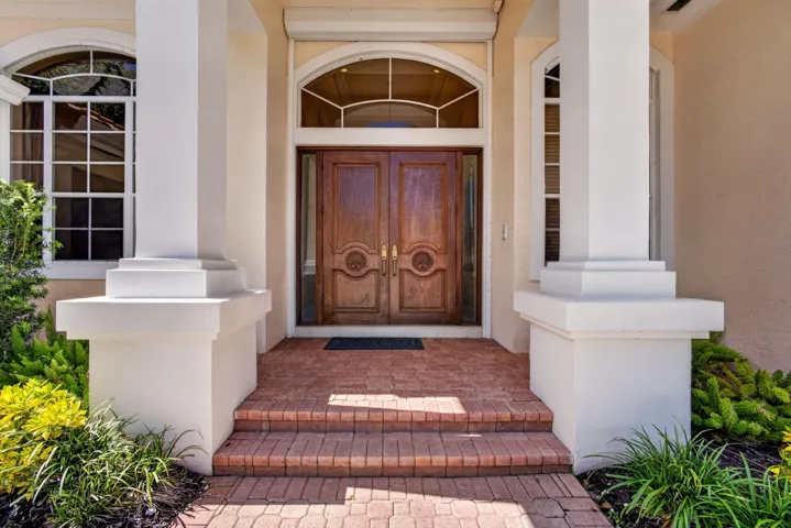 Double wood-paneled front doors with carved detail, arched transom window, brick masonry entry steps, and structural square columns