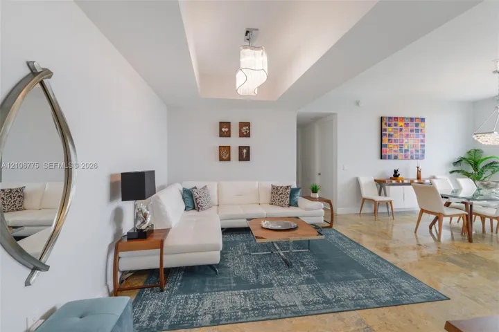 Open-concept room featuring a recessed tray ceiling, modern tiered light fixture, stone-finish flooring, and crisp white wall finishes