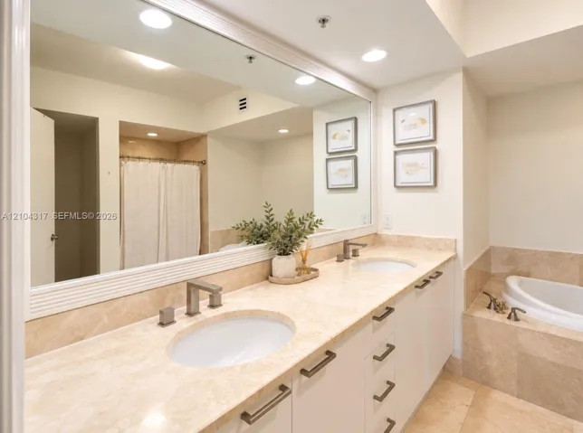 Dual-sink vanity with stone countertop, large-format mirror, recessed lighting, and built-in soaking tub with stone tile surround