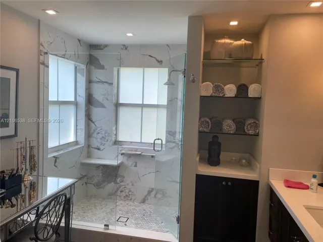 Walk-in shower featuring large-format patterned wall tile, a glass enclosure, a built-in stone bench, and recessed lighting, accompanied by built-in cabinetry with integrated shelving