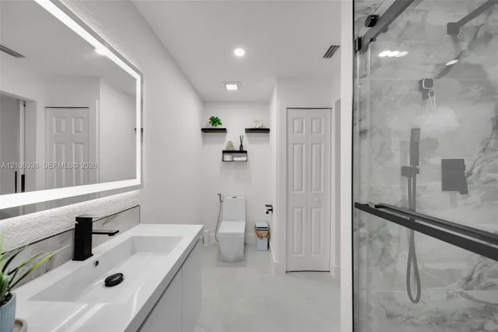 Modern vanity with integrated rectangular sink and matte black faucet, illuminated LED mirror, large-format tile flooring, walk-in shower with glass enclosure, and stone-finish wall tile