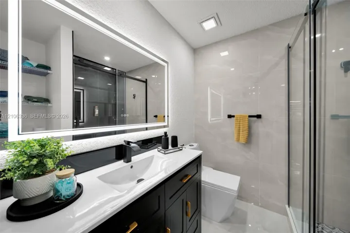 Bathroom vanity featuring a dark-finish cabinet with gold hardware, a white integrated basin, a polished countertop, an illuminated mirror, and a walk-in shower with large-format tile surround