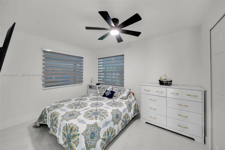 Neutral-toned bedroom featuring large-format tile flooring, a matte black multi-blade ceiling fan with integrated LED lighting, and dual windows equipped with horizontal zebra-style blinds