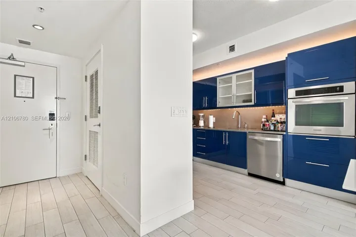 Modern kitchen featuring high-gloss blue cabinetry, built-in stainless steel oven, stainless steel dishwasher, under-cabinet lighting, and light-toned tile flooring