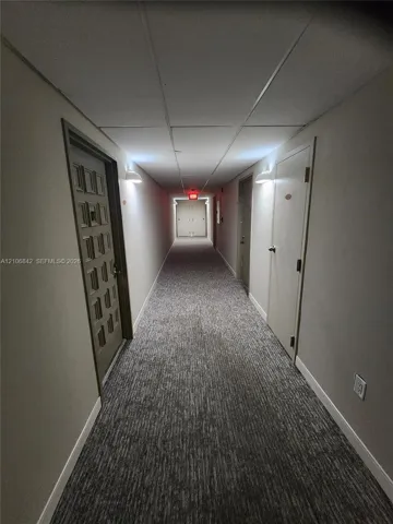 Interior hallway featuring neutral walls, grey patterned carpeting, textured drop ceiling tiles, and wall-mounted sconce lighting