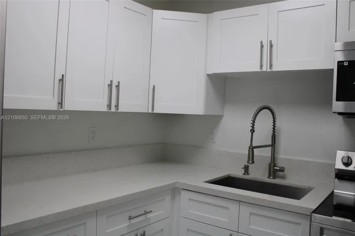 White Shaker-style cabinetry with polished metal hardware, light-toned solid surface countertops, a stainless steel undermount sink, and a high-arch pull-down faucet
