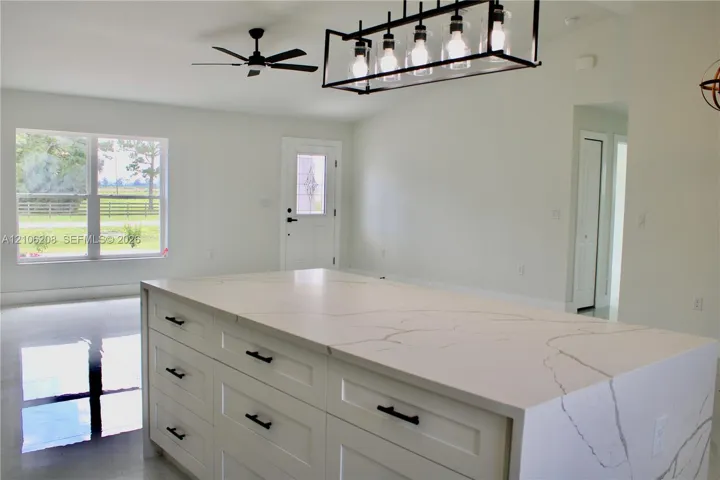 Large kitchen island featuring a veined stone countertop and white shaker-style cabinetry with matte black hardware, accented by a linear multi-light pendant fixture and a ceiling fan