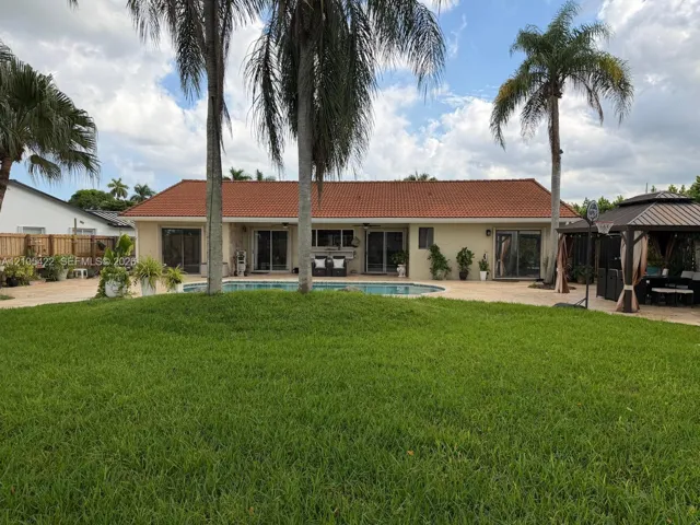 Expansive backyard featuring a large swimming pool, paver patio, permanent gazebo structure, and mature palm trees