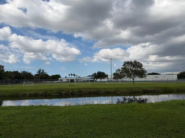 Open green space featuring a natural water element, perimeter fencing, and mature trees