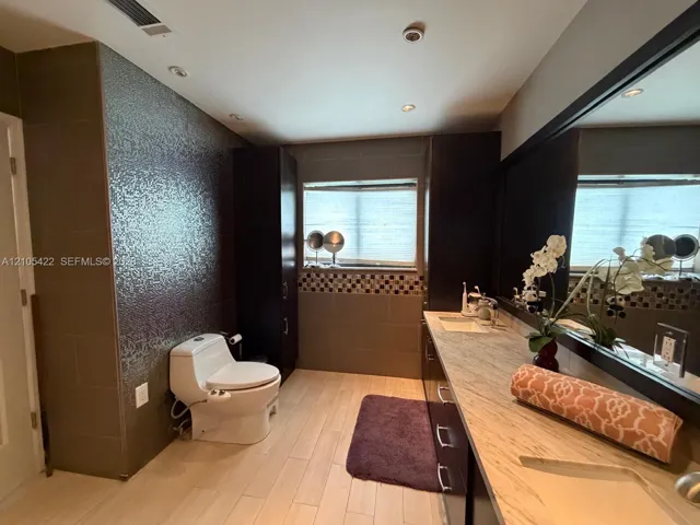 Bathroom featuring a stone-top vanity with dual integrated sinks, dark wood-finish cabinetry, floor-to-ceiling textured tile wall, and recessed ceiling lighting