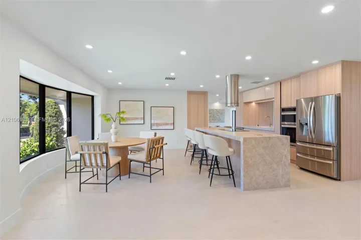 Modern kitchen featuring light wood-finish cabinetry, a large stone-surfaced island with a cylindrical exhaust hood, stainless steel appliances, recessed ceiling lighting, and expansive floor-to-ceiling windows