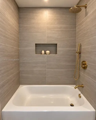 Bathtub with integrated shower featuring large-format tile surround, recessed wall niche, and brushed gold finish fixtures including a rainfall showerhead and handheld sprayer