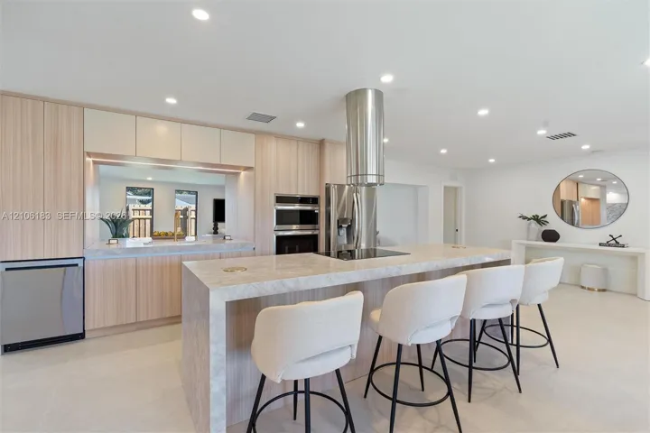 Contemporary kitchen featuring a large waterfall-edge island with light stone countertops, stainless steel appliances including a built-in double oven, polished concrete flooring, soft-close cabinetry in natural wood finish, and recessed ceiling lighting throughout the open-concept space