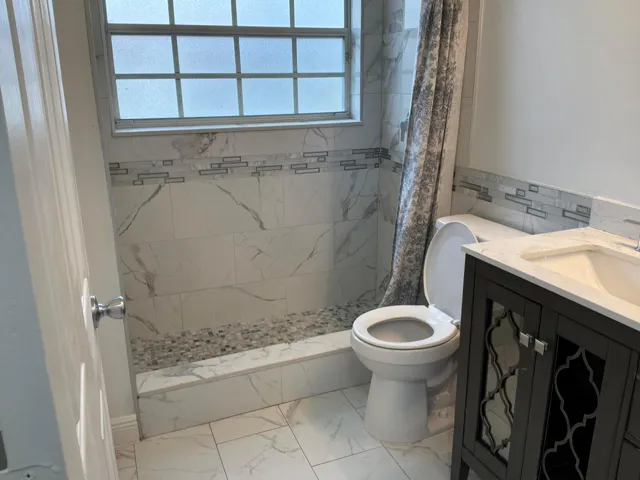 Bathroom featuring large-format light-toned tile flooring, dark cabinetry with glass-paneled doors, a white countertop, a shower area with decorative accent tiling, and a window with frosted​​‌​​​​‌​​‌‌​‌‌​​​‌‌​‌​‌​‌​​​‌​​ glass