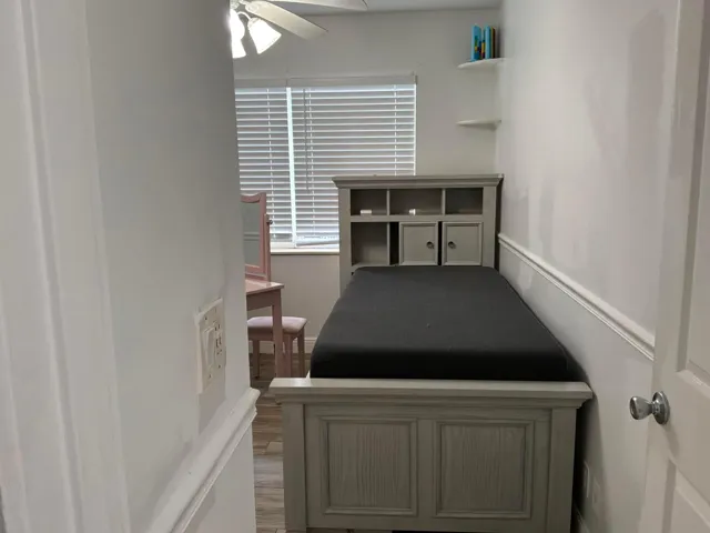 Wood-finish flooring, white horizontal window blinds, ceiling fan with integrated lighting, and wall-mounted floating shelves
