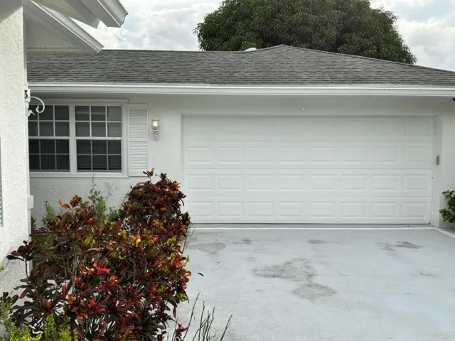 Exterior view featuring a white stucco facade, a multi-panel garage door, a shingled roof, a grid-patterned window, and exterior wall-mounted lighting