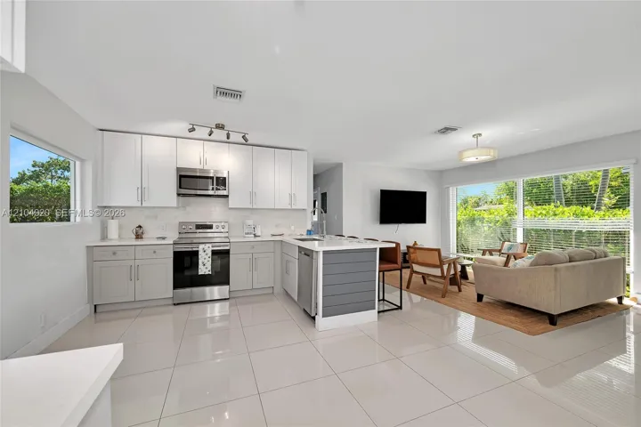 Kitchen featuring white shaker-style cabinetry, stainless steel appliances, a central island with a grey paneled base, quartz countertops, and large-format tile flooring