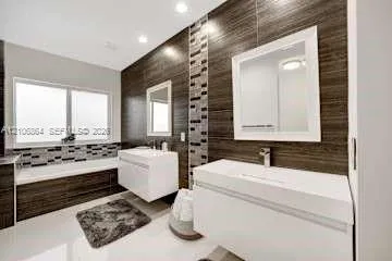 Dual floating vanities featuring white rectangular sinks and polished chrome faucets, complemented by horizontal wood-finish wall paneling, a glass mosaic tile accent strip, and a deep-soaking bathtub with a matching mosaic tile surround