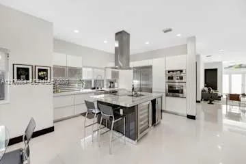 Modern kitchen featuring sleek flat-panel cabinetry, a central island with stone-finish countertops, stainless steel appliances, a chimney-style range hood, and polished flooring