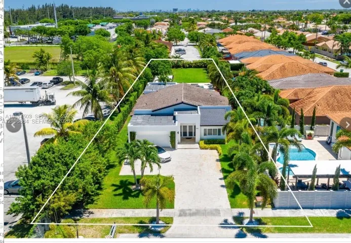 Aerial perspective featuring a residential property with a paved driveway, manicured lawn, mature palm trees, and an in-ground swimming pool