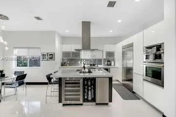 Kitchen featuring high-gloss white cabinetry, a center island with integrated wine refrigeration, stainless steel appliances including a built-in oven and microwave, and large-format tile flooring