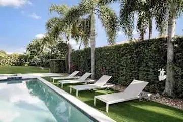 Rectangular swimming pool featuring a concrete deck and mature palm trees, bordered by a dense privacy hedge and synthetic turf area