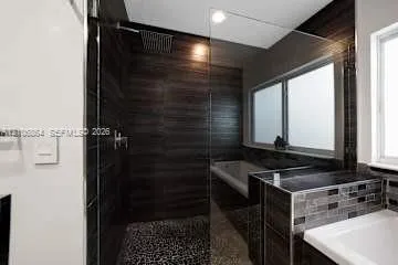 Walk-in shower with dark horizontal wall tile, pebble-texture floor, rain-style shower head, frameless glass enclosure, and a built-in soaking tub with mosaic tile surround