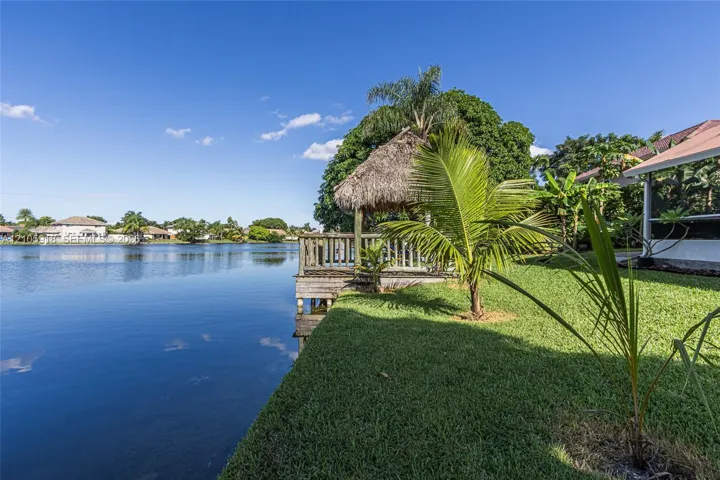 Waterfront backyard featuring a wooden deck with a thatched roof tiki hut, manicured lawn, and mature palm trees