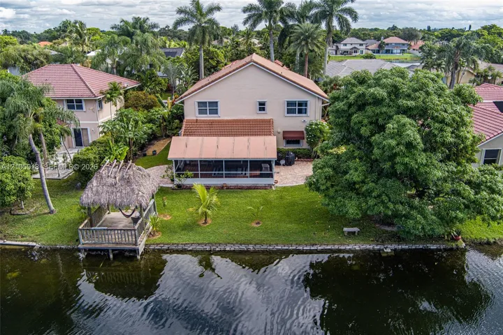 Waterfront two-story residence featuring a screened-in rear lanai, thatched-roof gazebo, paved patio area, and mature landscaping