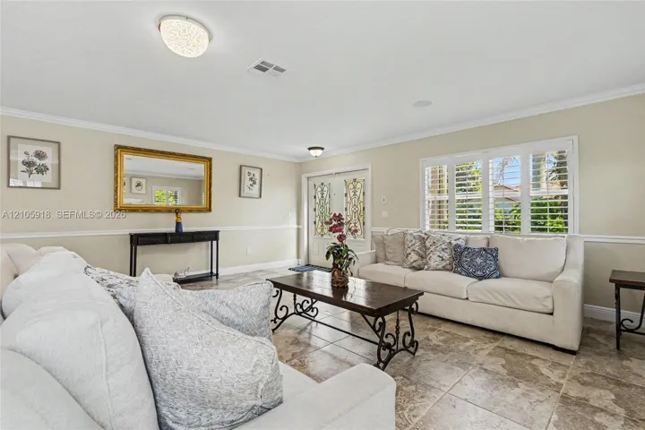 Neutral-toned interior featuring large-format tile flooring, crown molding, chair rail trim, dual entry doors with glass decorative inlays, and plantation shutters