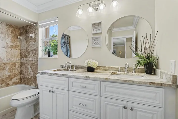 Bathroom featuring a dual-sink vanity with stone countertop, white shaker-style cabinetry, chrome fixtures, dual round wall mirrors, and tiled shower surround