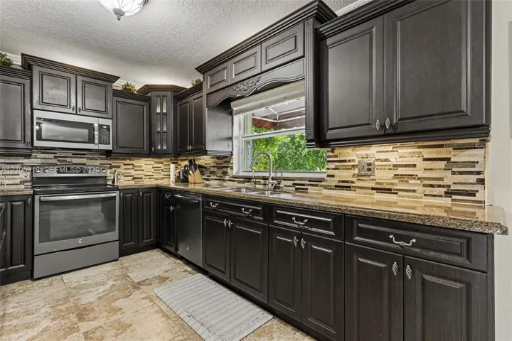 Kitchen featuring dark cabinetry with crown molding, stone-pattern tile flooring, multi-tonal linear backsplash, granite countertops, and stainless steel appliances