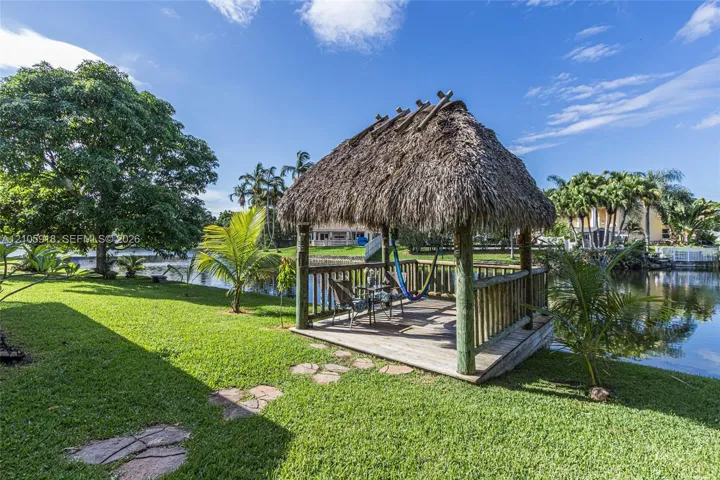Waterfront tiki-style gazebo featuring a thatched roof, timber structural posts, and wooden decking set upon a grass lawn with stone pavers