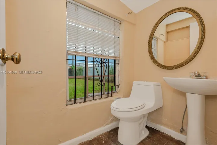 Window featuring iron security bars and horizontal blinds, pedestal sink, oval wall-mounted mirror, and low-profile toilet