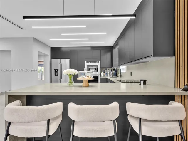 Modern kitchen featuring charcoal cabinetry, expansive stone-surface island, minimalist linear pendant lighting, built-in stainless steel double wall ovens, and vertical wood-slat accent wall detailing