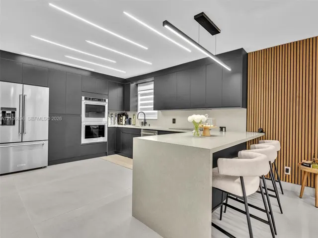 Modern kitchen featuring flat-panel cabinetry, a waterfall-edge stone island, linear LED ceiling lighting, integrated double wall ovens, and a vertical wood-slat accent wall