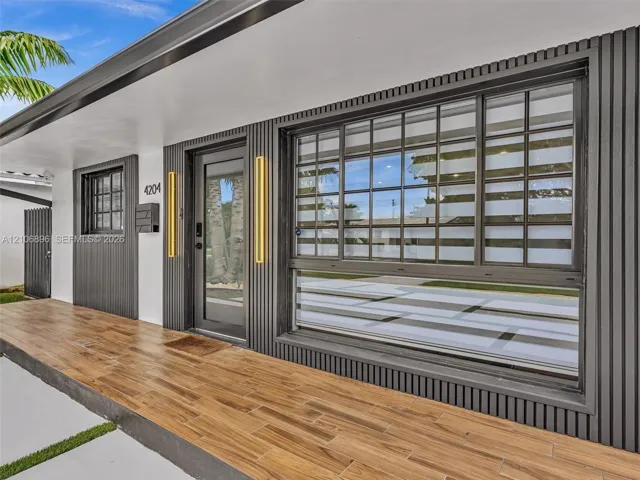 Exterior entry featuring wood-finish floor tiling, dark vertical slat siding, a glass-panel entry door, multi-pane sliding window, and dual vertical wall-mounted fixtures
