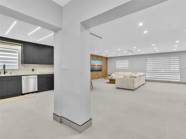 Open-concept area featuring large-format tile flooring, recessed ceiling lighting, kitchen cabinetry with a matte finish, wood-panel accent wall, and horizontal window blinds