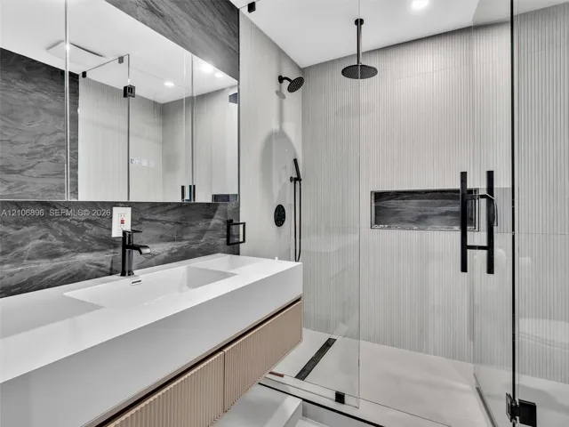 Floating vanity with an integrated sink and textured wood-finish cabinetry, paired with a frameless glass shower enclosure featuring matte black rainfall and handheld showerheads, a recessed wall niche, and large-format wall tile