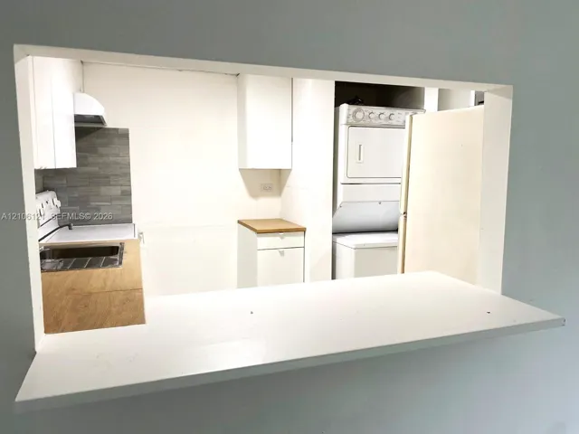 Kitchen pass-through window featuring a white ledge, light wood-finish cabinetry, gray tile backsplash, and a stacked laundry appliance configuration